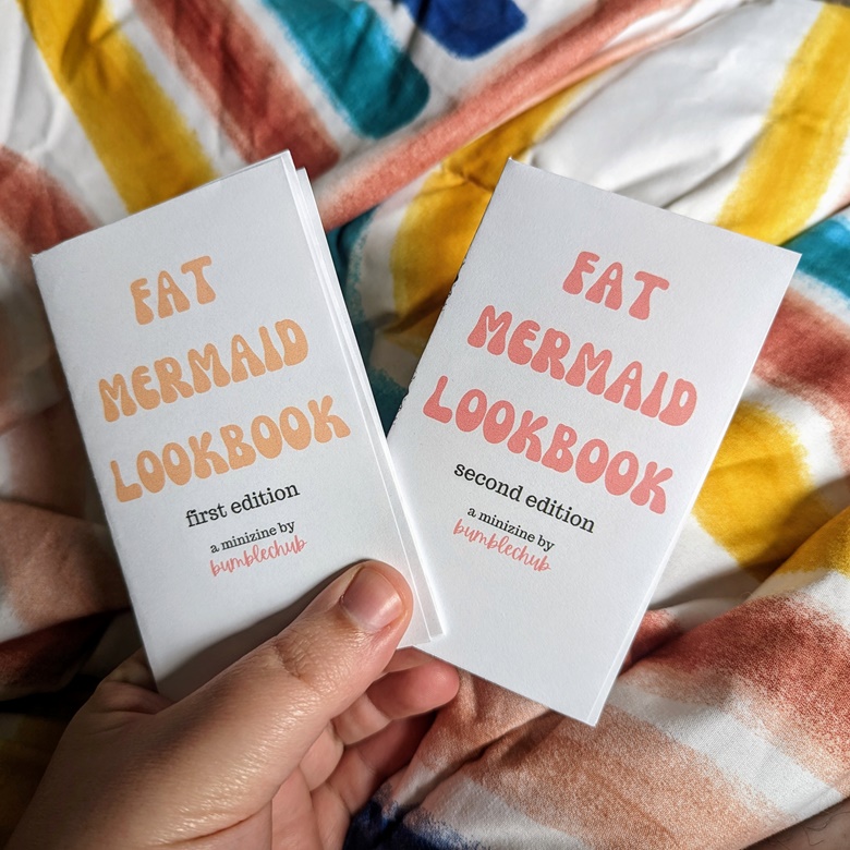  A white hand holding two minizines above a rainbow bedspread - the zines are "Fat Mermaid Lookbook" first and second editions, by bumblechub.