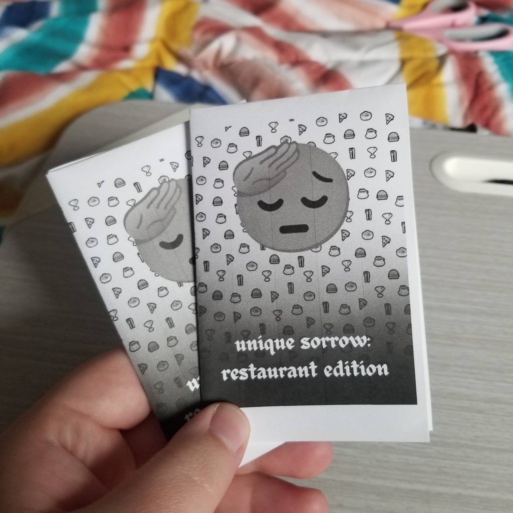  A white hand holding two black and white folded minizines. The cover reads “unique sorrow: restaurant edition” in a gothic, blackletter type font. In the background are spirals of food emojis - a coffee, martini, burger, pizza, etc. There is a large saluting face emoji in the center, looking sad.
